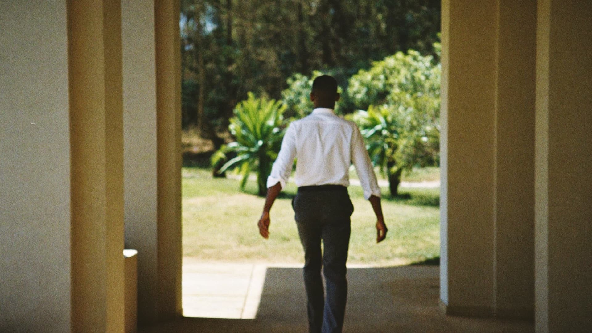 A person in a white shirt walking along a sunlit corridor towards a garden