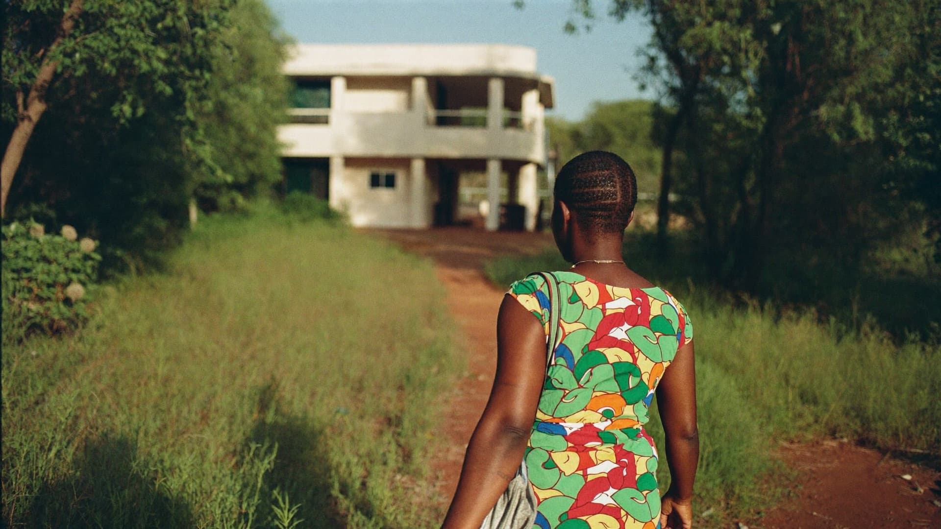 A woman seen from behind walking along a red-earth path towards a pale two-storey building, with trees and long grass on either side in late afternoon light