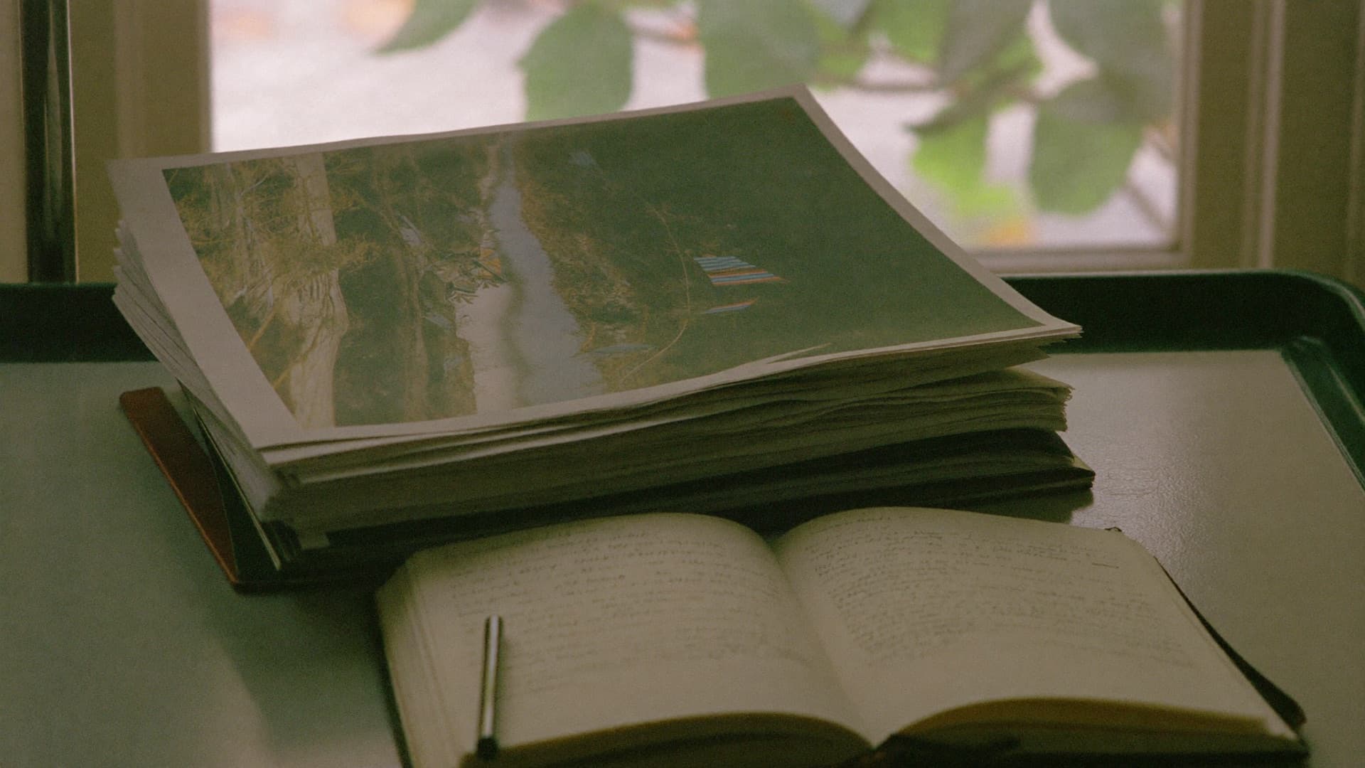 A stack of printed reports and an open notebook on a desk by a window